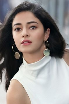 a woman with long black hair wearing large gold hoop earrings and a white turtle neck top