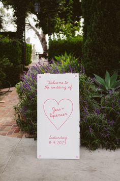 a welcome sign with a heart on it in front of some bushes and flowers at a wedding