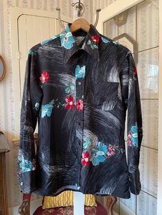 This beautiful shirt is made from a medium weight polyester blend in black, with a white and grey abstract pattern and dark red and teal blue flowers with green leaves. It has a pointy collar, the original buttons on the front and cuffs, and one chest pocket. The measurements, taken with the shirt lying flat, are: shoulder to shoulder, 19 inches; armpit to armpit, 23 inches; sleeves, 25 1/2 inches; length, 30 inches; bottom edge, 23 inches. In very good condition. Retro Black Button-up Blouse, Black Long Sleeve Tops With Abstract Print, Black Long Sleeve Top With Abstract Print, Black Floral Print Shirt For Summer, Spring Patterned Shirt With Bold Print, Fitted Shirt With Abstract Print For Spring, Vintage Black Shirt With Button Closure, Vintage Black Button-up Blouse, Black Collared Top With Floral Print