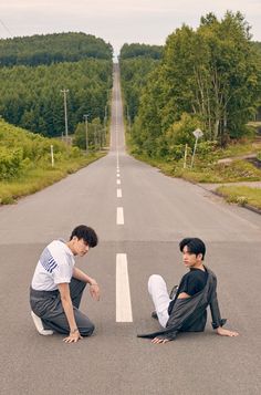 two young men sitting on the side of an empty road with trees in the background