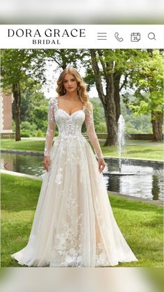 a woman wearing a wedding dress with long sleeves and flowers on the skirt is standing in front of a pond