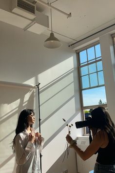 two women standing in front of a window and one is filming another woman with a camera