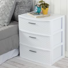 a white nightstand with two drawers next to a bed and a plant on top of it