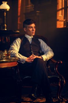 a man sitting in a chair wearing a vest and tie with his hands folded out