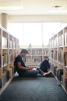 a person sitting on the floor in front of a book shelf with many books and looking at a laptop