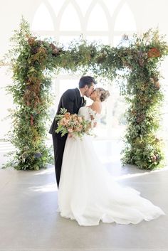 a bride and groom standing in front of an archway with greenery on it at their wedding