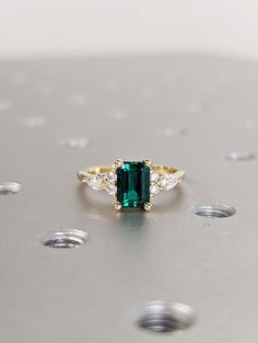 an emerald and diamond ring sitting on top of a silver surface with small white dots around it