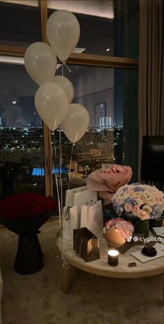 balloons and flowers on a table in front of a window with the city lights behind them