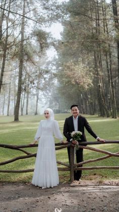 a man and woman standing next to each other in front of a fence with trees