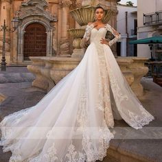 a woman standing in front of a fountain wearing a wedding dress with long sleeves and lace detailing