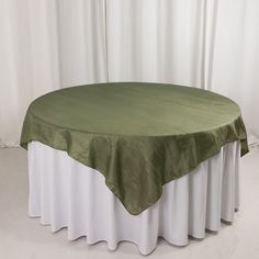 a round table covered with a green and white linen over it's top, in front of a curtained window