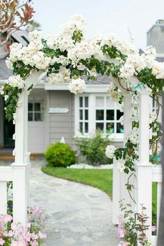 a white arbor with flowers and greenery on the side of it in front of a house