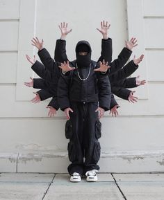 a person with their hands in the air and wearing a black outfit, standing next to a white wall