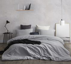 a bed with grey sheets and pillows in a white room next to a book shelf