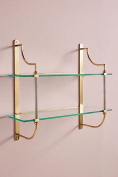 two glass shelves on the wall with brass brackets and hooks attached to them, against a pink wall