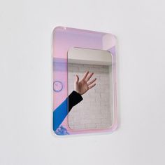 a person's hand reaching out from behind a mirror on a white wall with pink and blue background