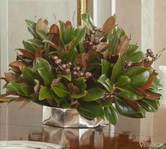 a vase filled with lots of green plants on top of a wooden table next to a mirror