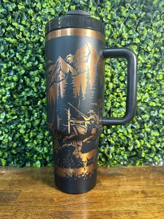 a black and gold travel mug sitting on top of a wooden table next to a green bush