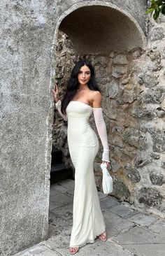 a woman in a white dress is standing by a stone wall and posing for the camera