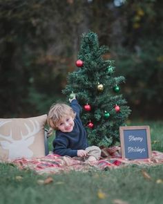 a little boy sitting on a blanket in front of a christmas tree with a chalkboard sign