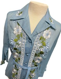 Really pretty vintage button up shirt with embroidered flowers on front, collar, and sleeves, and fabric covered buttons. Made of (I believe) a sheer muslin fabric with a nice sheen. Shirt has a matching fabric belt. Dates to around the 1970s and possibly made in China or Japan. Long sleeves widen at the wrist. Was found in the garage of an estate sale. It was hanging on a metal hanger and was very dirty. It cleaned up very well but does has a very faint rust stain remaining on shoulder and inside near collar. Please see photos with arrows. No holes or rips. The only label is the for the size. I'd say it fits like a medium but please FOLLOW MEASUREMENTS BELOW for the best fit. Would be beautiful paired with a white skirt or slacks or even jeans. **Please refer to the following measurements Fitted Embroidered Button-up Shirt, Blue Embroidered Collared Blouse, Spring Button-up Blouse With Embroidered Cuffs, Spring Embroidered Cuffs Button-up Blouse, Vintage Blue Embroidered Blouse, Spring Shirt With Embroidered Spread Collar, Spring Shirt With Embroidery And Spread Collar, Spring Embroidered Shirt With Spread Collar, Fitted Embroidered Collared Shirt