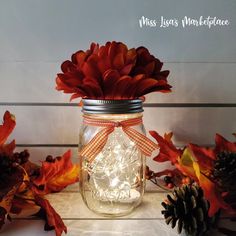 there is a mason jar with lights in it next to some leaves and pine cones