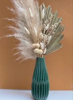 a green vase filled with lots of white flowers and dry grass on top of a table