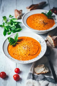 two bowls of tomato soup with bread and basil on the side, next to tomatoes