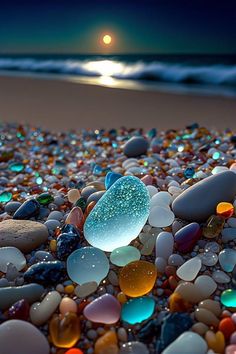 the beach is covered in many different colored rocks and pebbles at night, with a full moon rising over the horizon