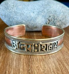 "This beautiful Om Namah Shivaya cuff bracelet is made in Nepal. Size: 18cm - As It's open-ended, would fit most hands Width - 15mm approx Weight - 23gms approx Material: white metal, brass, copper To fit the bracelet open it up so the gap is wide enough to pass over the narrowest part of your wrist and then squeeze it back into shape until it is a nice comfortable fit. Om Namah Shivaya (Devanagari: ॐ नमः शिवाय;[1] IAST: Om Namaḥ Śivāya) is one of the most popular Hindu mantras and the most impo Handmade Adjustable Spiritual Bangle, Symbolic Handmade Bracelets For Festivals, Adjustable Symbolic Bangle Bracelet, Handmade Spiritual Cuff Bracelet For Festivals, Spiritual Festival Bangle Cuff Bracelet, Handmade Adjustable Bangle For Festival, Spiritual Silver Brass Bracelets, Silver Brass Spiritual Bracelets, Handmade Symbolic Beaded Bangle Bracelet