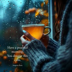 a woman holding a cup of tea in front of a window with rain drops on it