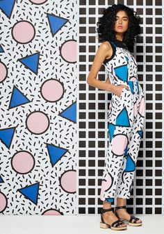 a woman standing in front of a wall with geometric designs on it and wearing sandals