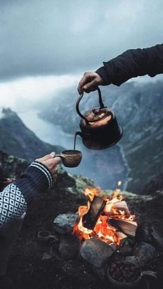 two people holding tea kettles over an open fire with mountains in the background on a cloudy day