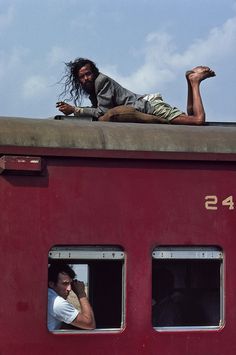 a man sitting on top of a red train next to another man with long hair