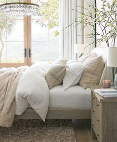 a bed with white sheets and pillows next to a lamp on a table in front of a window