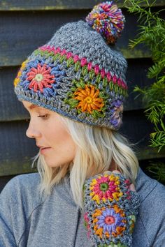 a woman wearing a knitted hat and mitten with flowers on it, standing in front of a wooden fence