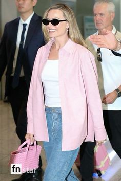 a woman in pink jacket and jeans walking through an airport with her hand on her purse