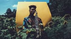 a man holding a saxophone in front of a large yellow object with trees and bushes behind him