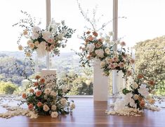 three tall white vases with flowers on top of a wooden table in front of large windows