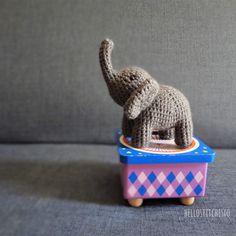 a knitted toy elephant standing on top of a blue and pink box with its trunk in it's mouth