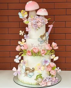 a three tiered cake with flowers and butterflies on it's side, sitting in front of a brick wall