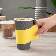 a hand holding a yellow paper cup with coffee in it on top of a wooden table