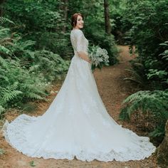 a woman in a wedding dress standing on a dirt path surrounded by trees and ferns