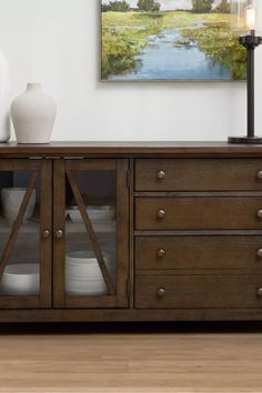 a wooden cabinet with glass doors and plates on the bottom, in front of a painting