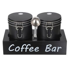 two black coffee mugs and spoon on a wooden stand with the words coffee bar painted on it