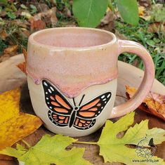 a ceramic mug with a butterfly painted on it sitting next to some leaves and grass