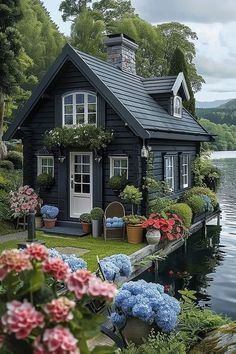 a black house with flowers and potted plants in front of it next to the water