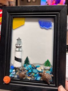 there is a picture frame made out of sea glass and rocks with a lighthouse in the background