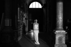 black and white photo of woman in long dress walking through an old building with columns