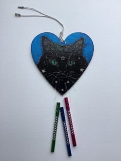 an ornament shaped like a heart with a black cat on it and three colored pencils next to it